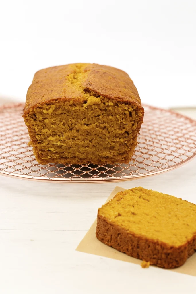 Vertical imaVertical image of pumpkin bread on a copper cooling rack with a sliced piece on parchment paper, styled for rustic food photography.ge of pumpkin bread on a copper cooling rack with a sliced piece on parchment paper, styled for rustic food photography.