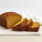 Sliced pumpkin bread with golden crust and orange interior on a white marble board with soft cloth background.