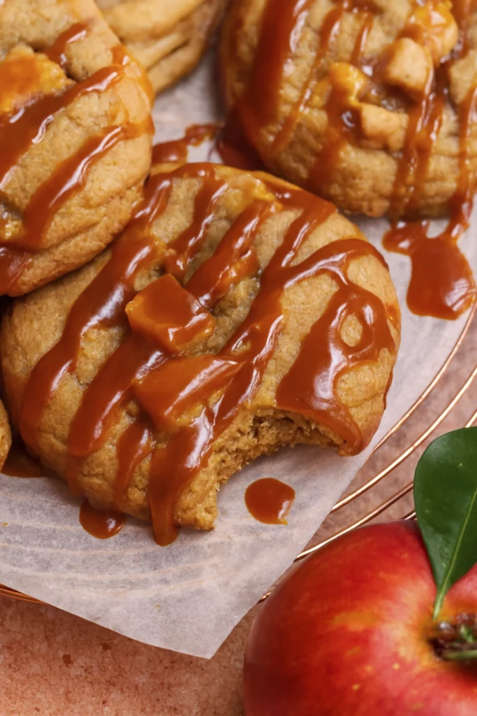 Caramel apple cookies with apple chunks and caramel drizzle on a parchment-lined cooling rack, styled with a fresh red apple for autumn dessert appeal.