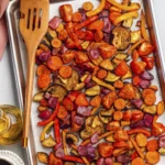 Colorful roasted vegetables including carrots, peppers, zucchini, eggplant, and onions on a baking sheet with olive oil and balsamic vinegar