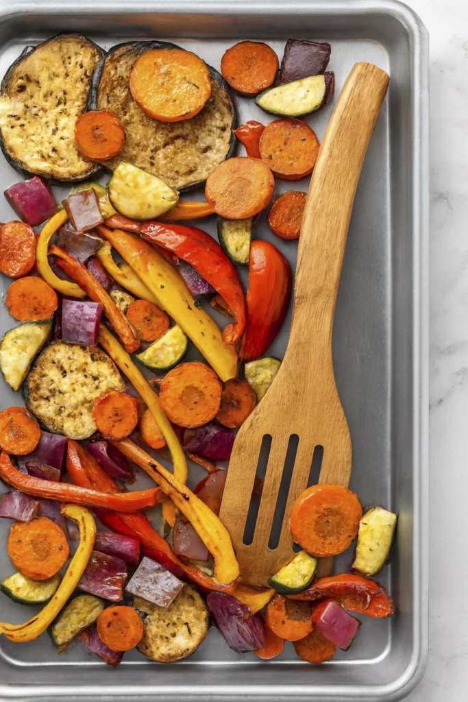 Rustic tray of roasted vegetables including carrots, peppers, zucchini, eggplant, and onions with a wooden spatula