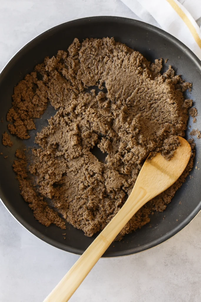 Cooked ground beef in black frying pan with wooden spatula on light surface and striped cloth