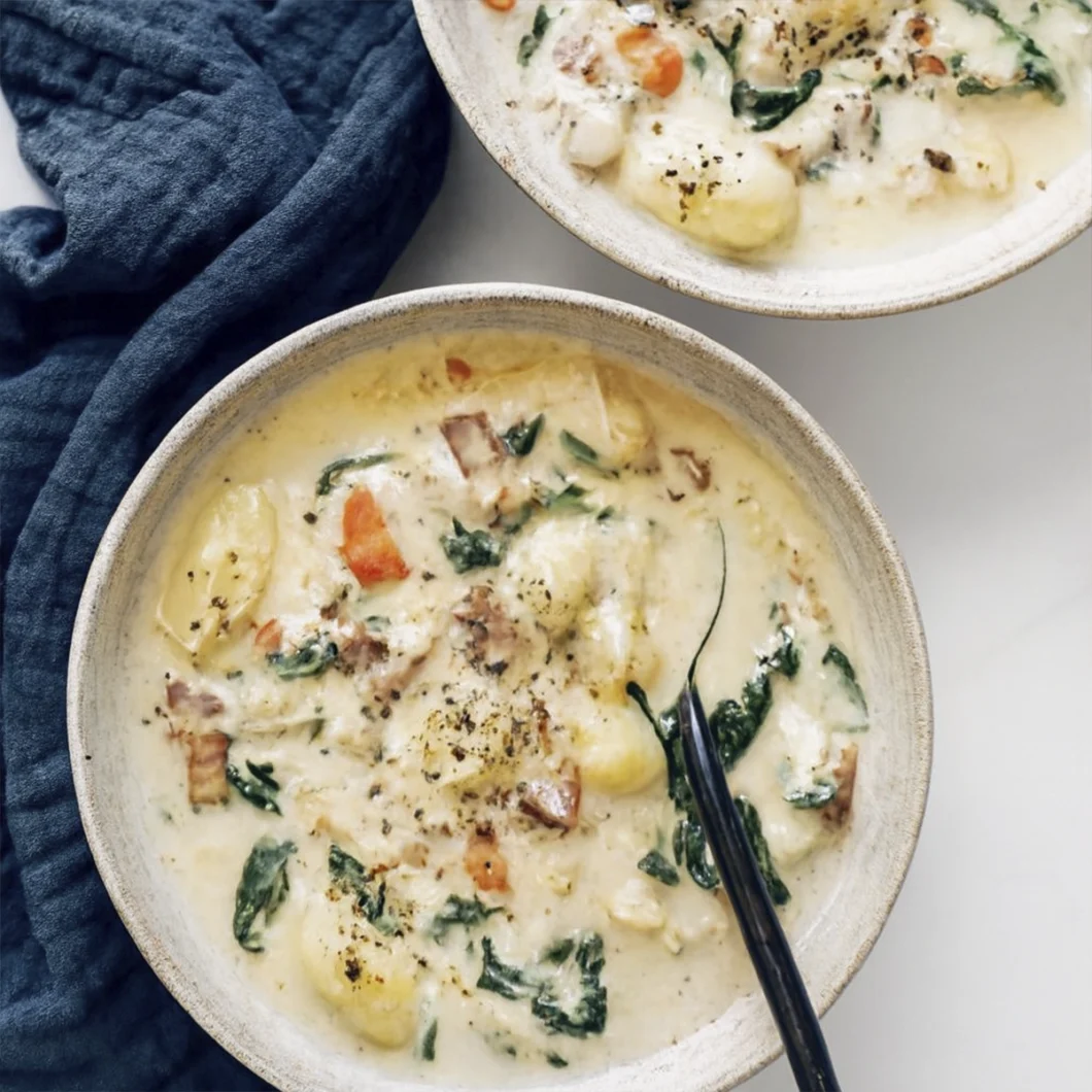 Two bowls of creamy gnocchi soup with spinach, carrots, bacon, and chicken on a white surface.