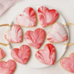 Heart-shaped cookies with pink marbled icing on a white plate with woven handles.
