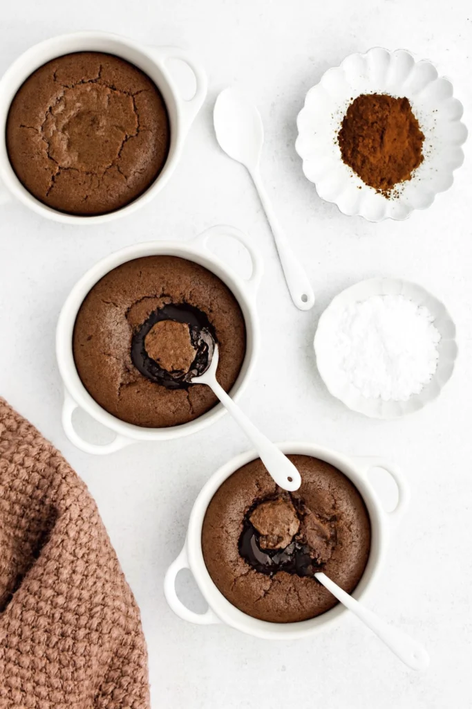 Gooey chocolate lava cakes in white ramekins on marble with powdered sugar and cocoa styling.