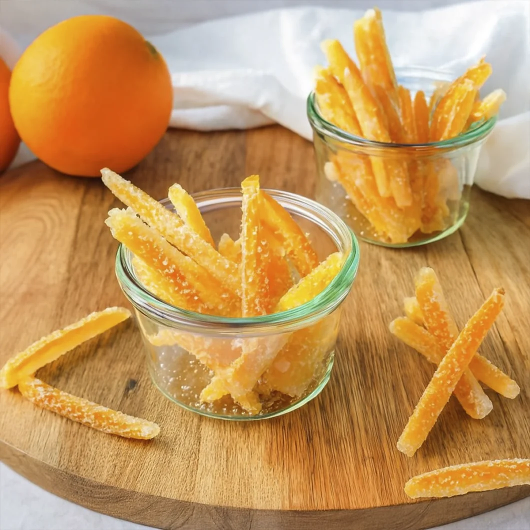 Glass jars filled with sugar-coated candied orange peels on a rustic wooden board with whole oranges in the background.