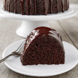 Moist chocolate sour cream bundt cake with glossy glaze on a white cake stand and plated slice