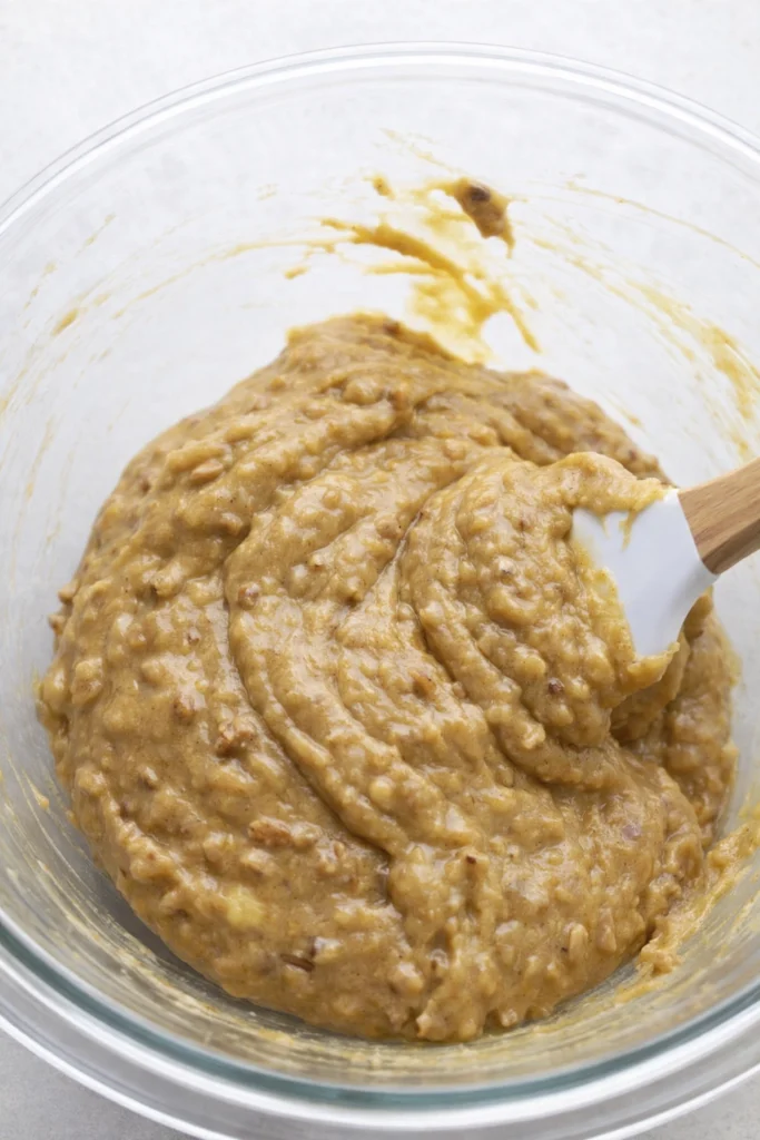 Thick banana bread batter with nuts in a glass bowl, stirred with a spatula.