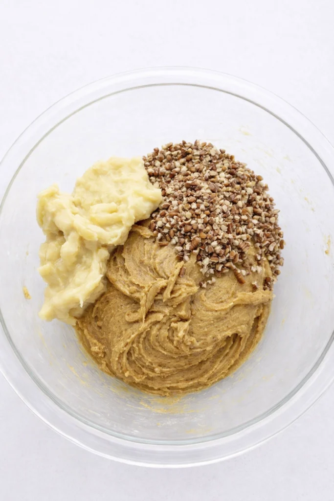 Glass mixing bowl with brown batter, mashed bananas, and chopped nuts ready to be combined.