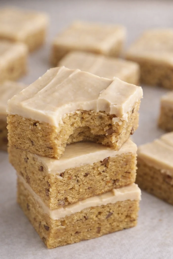 Stack of frosted blondie bars with creamy beige frosting, one bar showing a bite.