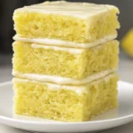 Close-up of three stacked lemon dessert bars with glossy glaze on a white plate, showcasing moist texture and bright kitchen lighting.