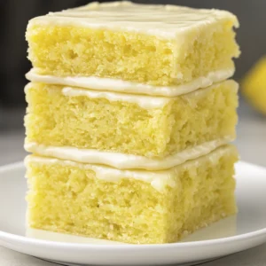 Close-up of three stacked lemon dessert bars with glossy glaze on a white plate, showcasing moist texture and bright kitchen lighting.
