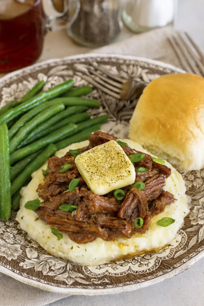 Plated Mississippi pot roast with shredded beef over mashed potatoes, butter, green onions, green beans, and a dinner roll on a floral dish