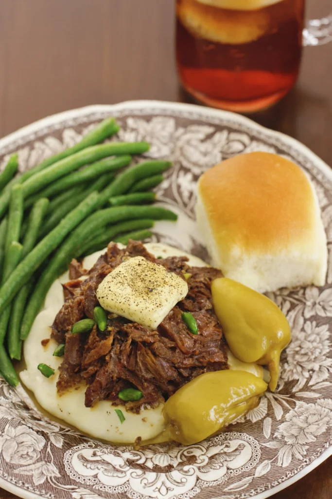 Mississippi pot roast served over mashed potatoes with butter and garnish, whole yellow peppers, green beans, dinner roll, and iced tea with lemon