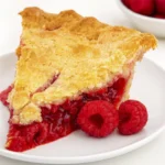 Slice of raspberry pie with golden crust and glossy red filling on a white plate with fresh raspberries