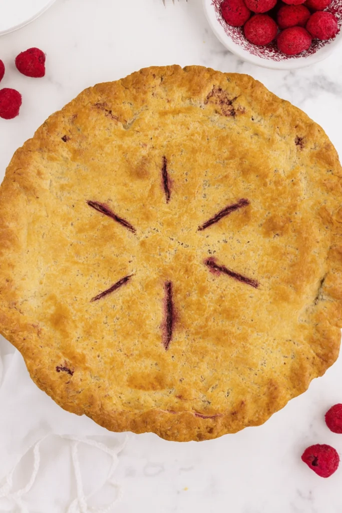 Whole raspberry pie with golden crust and six slits, surrounded by fresh raspberries and vintage forks