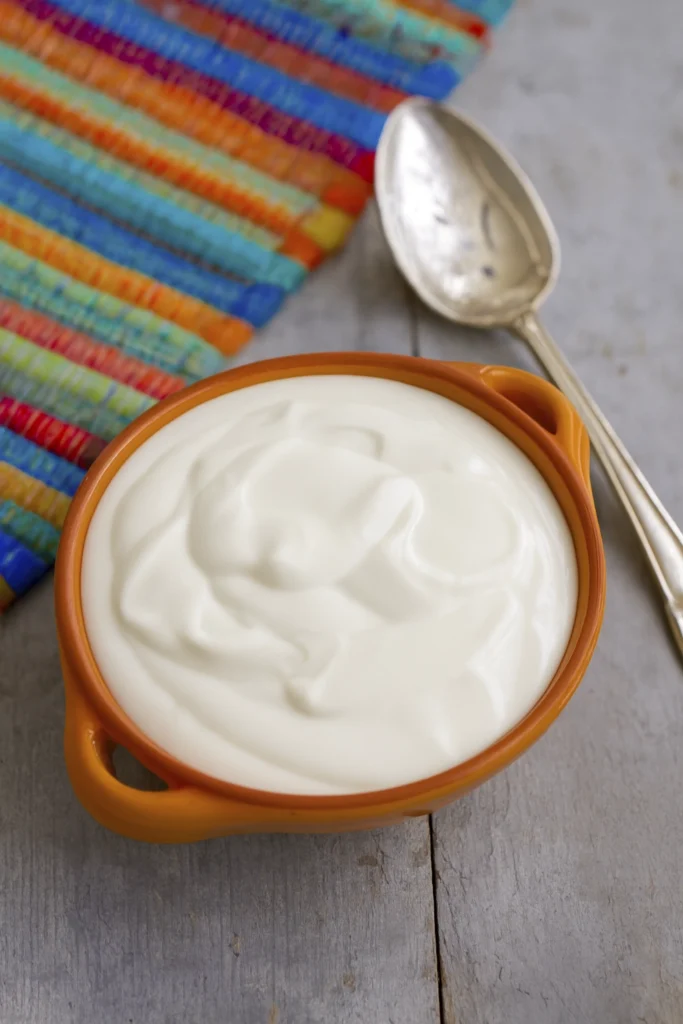 Thick creamy sour cream in an orange ceramic bowl on rustic wood with colorful woven cloth in background