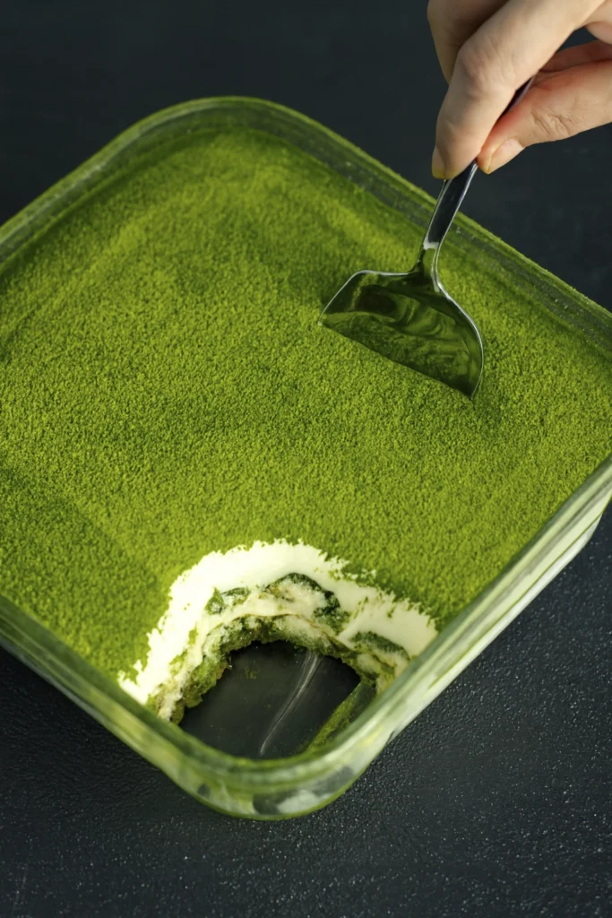 Square glass dish of matcha tiramisu with creamy layers and matcha powder topping, being served with a utensil.