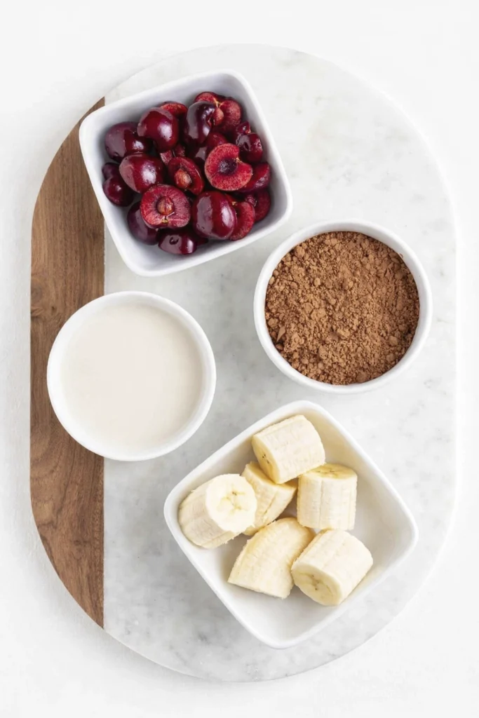 Fresh cherries, cocoa powder, banana slices, and milk in white bowls on a cutting board.