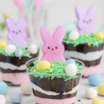 Easter dirt cups with pink cream, cookie crumbs, green coconut grass, marshmallow bunnies, and pastel candy eggs.