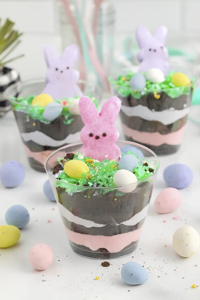 Elegant Easter dirt cups with cream layers, cookie crumbs, coconut grass, sprinkles, candy eggs, and marshmallow bunny Peeps.