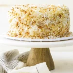 Close-up of a moist coconut cake slice with creamy frosting and toasted coconut flakes on a white plate with a gold fork.