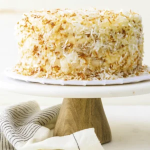 Close-up of a moist coconut cake slice with creamy frosting and toasted coconut flakes on a white plate with a gold fork.