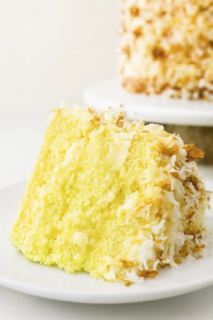 Coconut cake slice with creamy frosting and toasted coconut flakes on a white plate with gold fork.